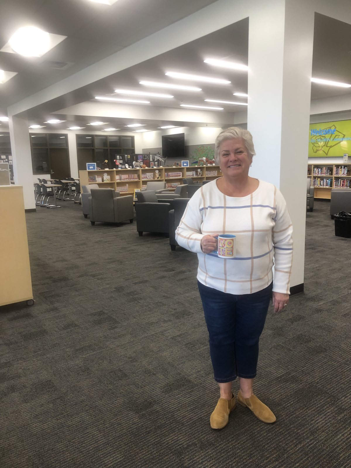 Colleen Martin holding a mug that reads VOTE inside a modern school library commons