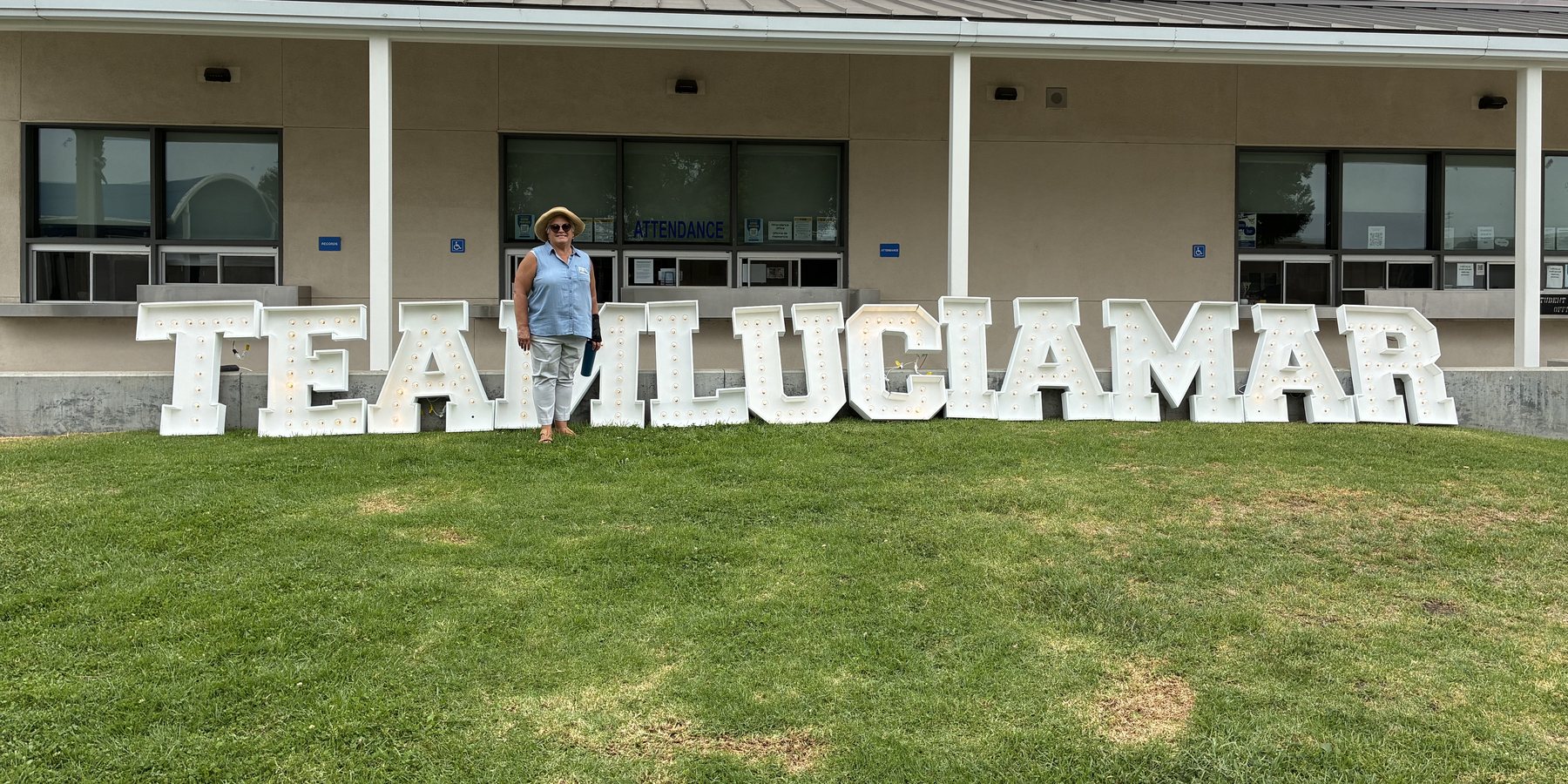 Colleen Martin standing in front of large illuminated letters spelling TEAM LUCIA MAR outside a district school